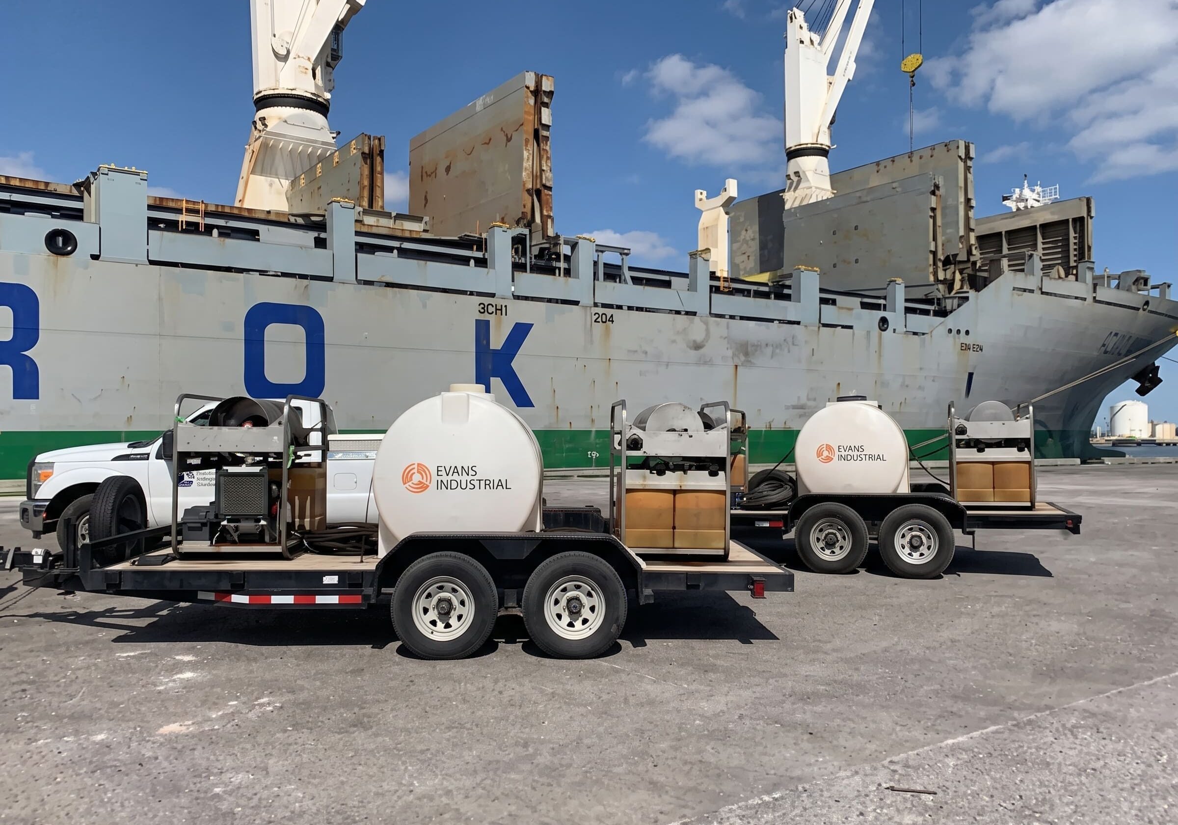 Two Evans Industrial trailer-mounted hot water pressure washers with water tanks and hose reels parked on a port dock beside a cargo ship