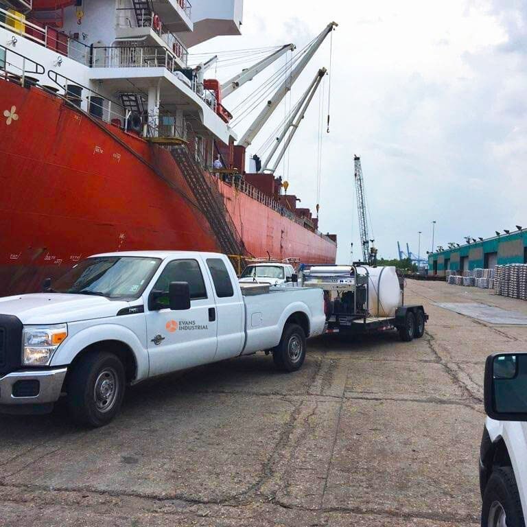 Evans Equipment truck towing a pressure washer trailer along a port dock beside a cargo ship