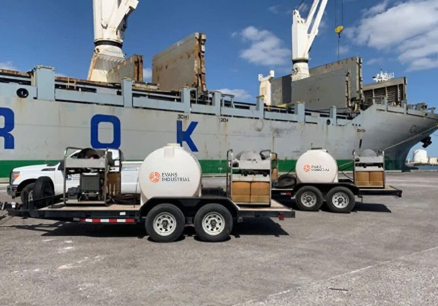 Two trailer-mounted hot water pressure washers with water tanks parked on a port dock beside a cargo ship